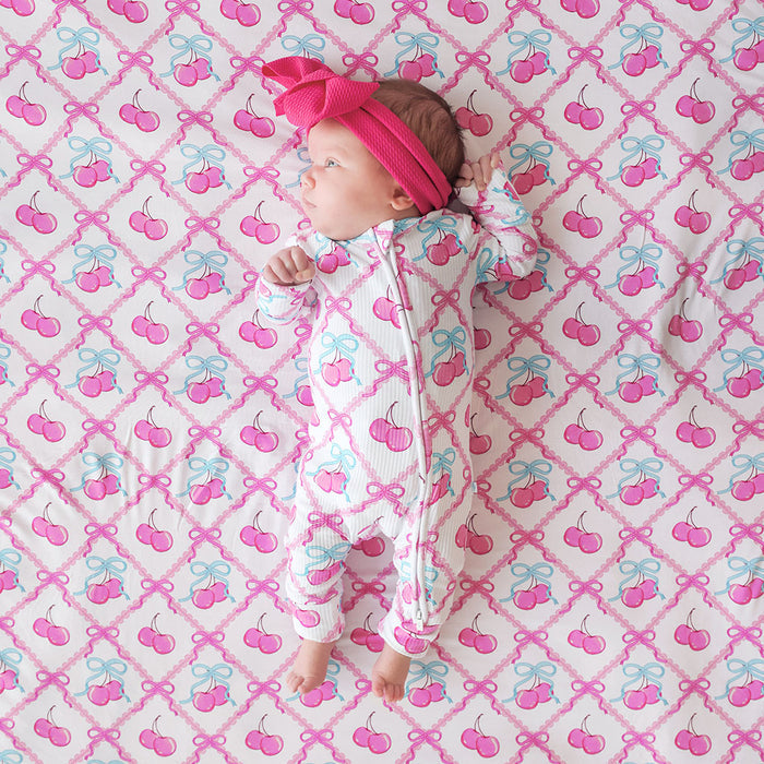 Baby in a cherry-patterned one piece lying on a pink and blue bow and cherry patterned blanket.