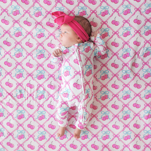 Baby in a cherry-patterned one piece lying on a pink and blue bow and cherry patterned blanket.