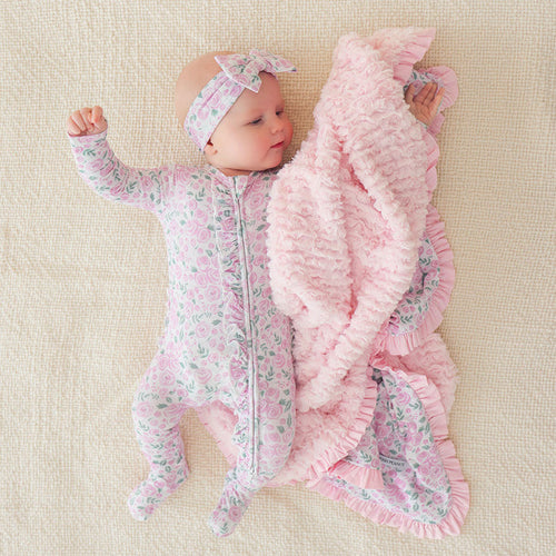 infant laying down on beige colored sheet wearing floral themed print one piece with matching bow headwrap and plush blanket