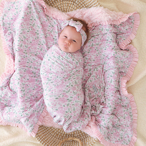 Baby wrapped in a pink floral swaddle blanket with matching headband with ruffles on a beige surface
