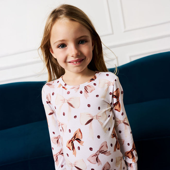 Young girl wearing a brown and white coffee beans and bows printed pajama set against a white wall.