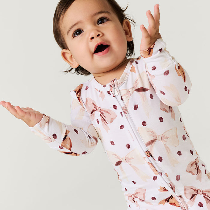 Baby wearing a white long-sleeved one piece with brown bows and coffee beans pattern on a light gray background