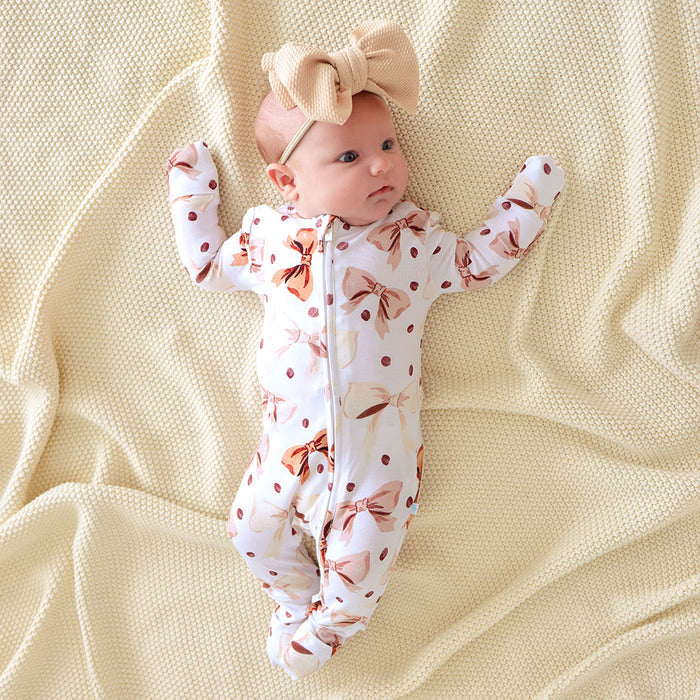 Baby wearing a white and brown coffee beans and bows one piece laying a beige textured blanket.