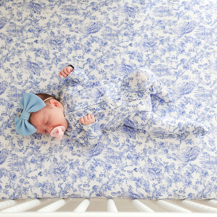 Newborn baby wearing a blue printed one piece lying on a blue floral crib sheet.