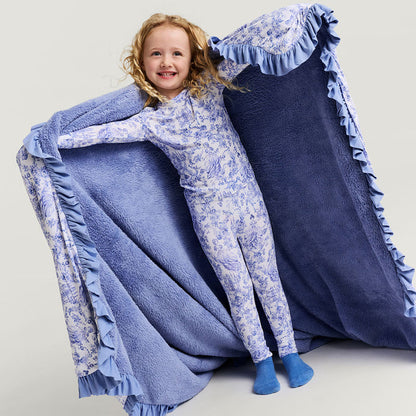 Child in a floral onesie standing next to a large blue blanket with ruffles on a light gray background