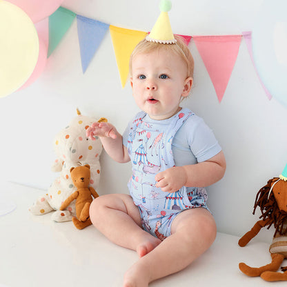 Baby sitting wearing circus printed french terry short overalls next to stuffed animals.
