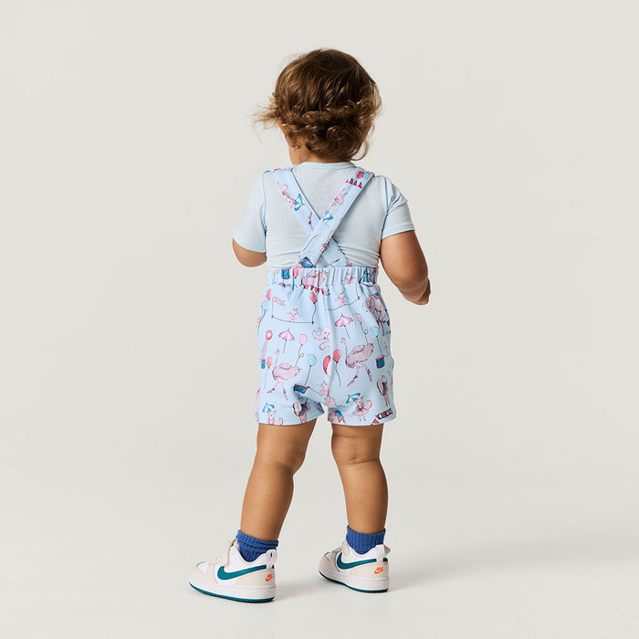 back image of baby standing wearing circus printed short overalls.