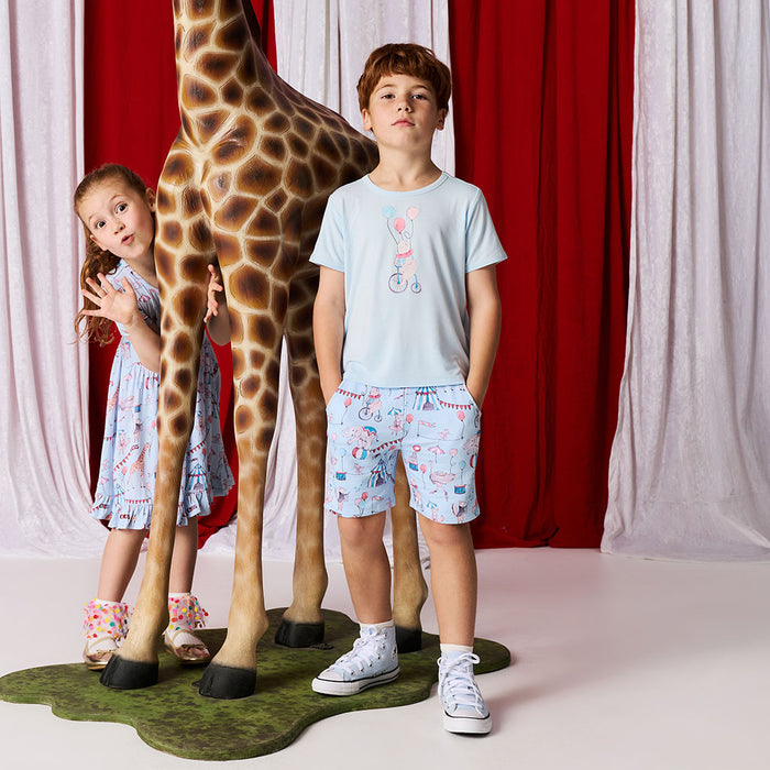 Two children standing next to a life-size giraffe prop with a red and white curtain background.