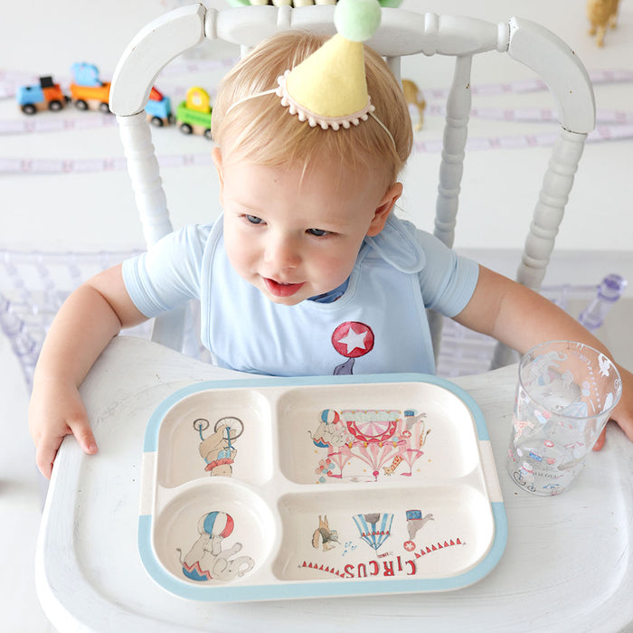 Baby sitting in a high chair with a tray featuring circus designs, surrounded by toys and a glass.