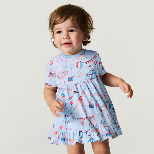 Child wearing a light blue dress with colorful circus-themed print on a plain background