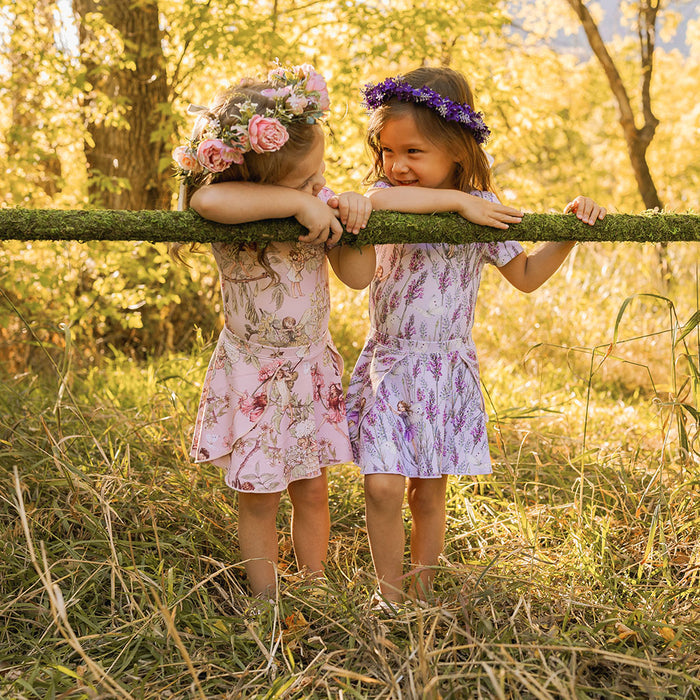 Two young girls in floral dresses and flower crowns standing behind a wooden fence in a sunlit forest.