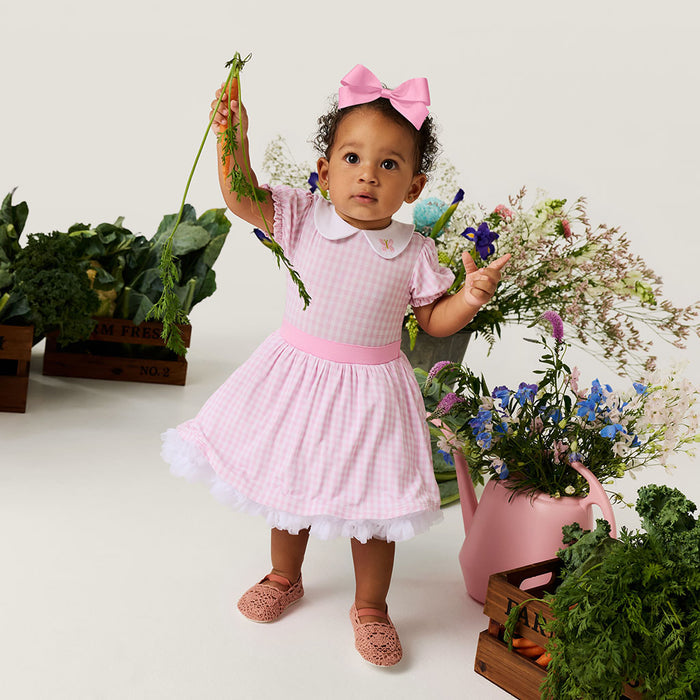 Child in a pink petticoat dress holding flowers and carrots, surrounded by plants and flowers.