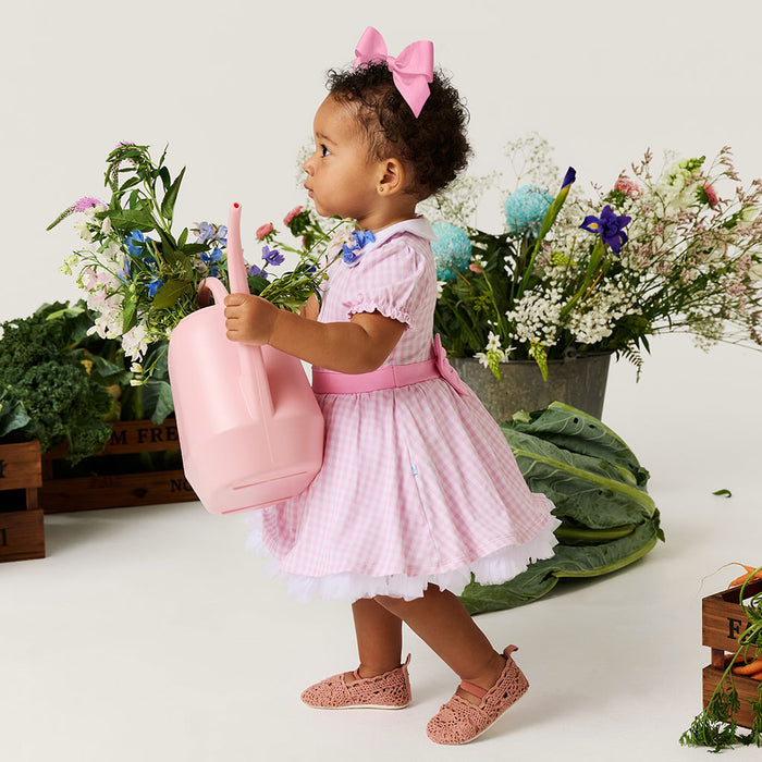 Child in a pink dress holding a pink watering can with flowers and plants in the background