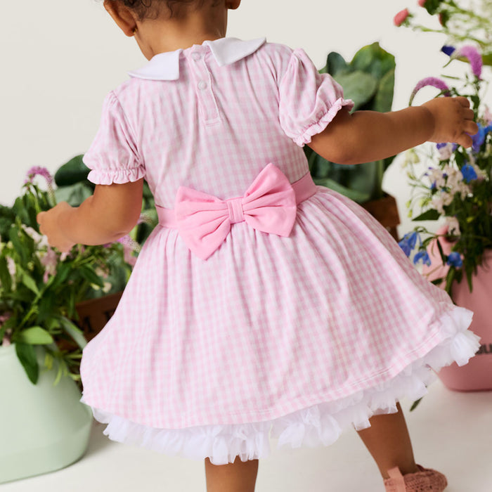 Child wearing a pink checkered dress with a large bow, standing among potted plants.