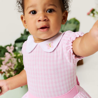 Baby wearing a pink checkered dress with a white collar, standing against a light background.