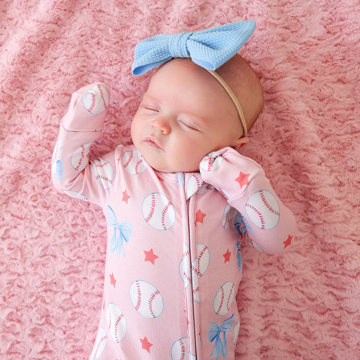Newborn baby in a pink outfit with baseball-themed design, wearing a blue bow headband, on a pink blanket.
