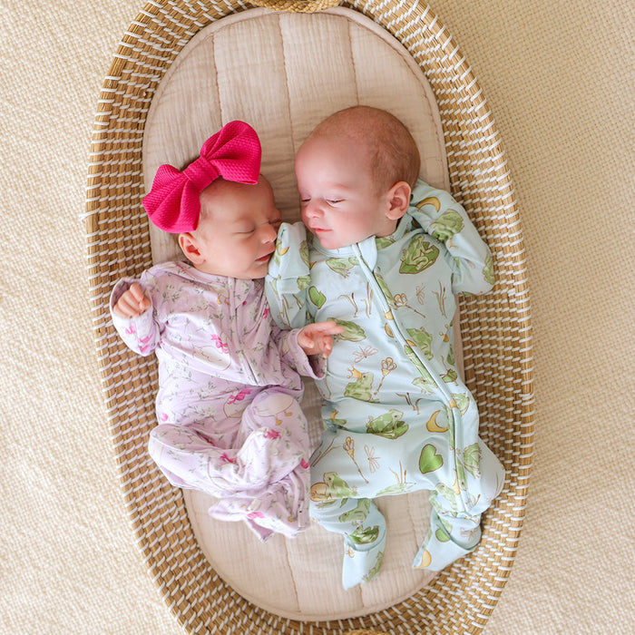 Two babies in a woven basket, one wearing a pink bow and the other in a green and white outfit wearing bamboo viscose pink and green one pieces.