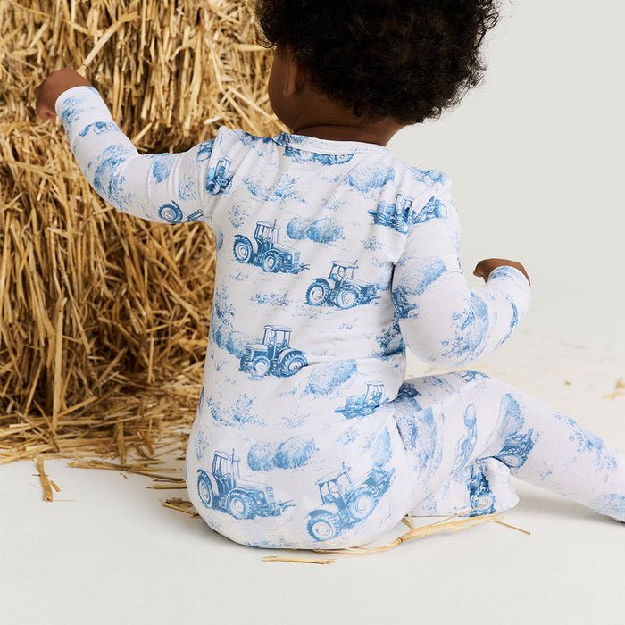 Child wearing a white onesie with blue tractor pattern, sitting next to a bale of hay.