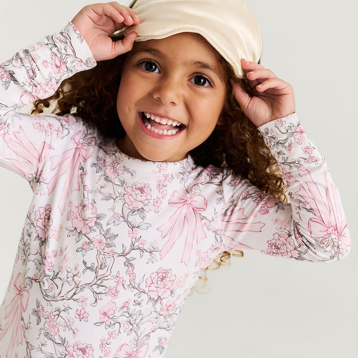 close up of child wearing a floral and bow printed pajama set holding a sleeping mask over head