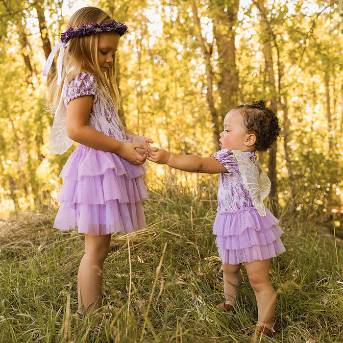 Two young girls in matching purple dresses standing in a forest during sunset.