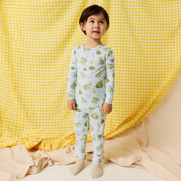 Young boy  wearing blue bamboo viscose pajama set with frog prince print standing in front of a yellow checkered background.