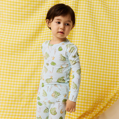 Young boy wearing blue bamboo viscose pajama set with frog prince print standing in front of a yellow checkered background.