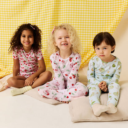 Three children wearing pajama sets with strawberry, mouse, and frog prince prints sitting in front of a yellow checkered background.