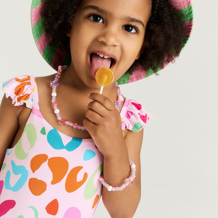 Child wearing a colorful jumpsuit and hat, holding a lollipop against a plain background