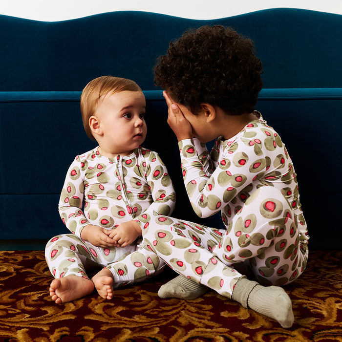 Two children in matching olive pajamas sitting on a patterned rug with a blue couch in the background.