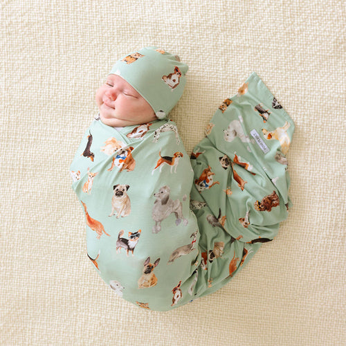 Newborn baby swaddled in a green blanket with matching beanie and dog pattern on a beige surface