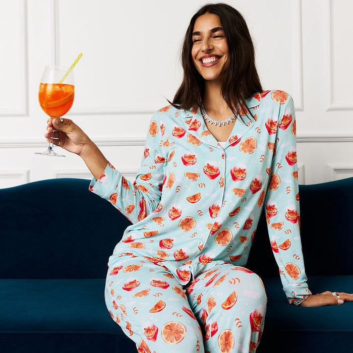 Women sitting on blue coach holding a cocktail wearing a aperol spritz printed pajama set.