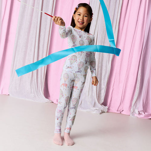 Child wearing a pajama set with circus animal and hot air balloon print waving a ribbon standing against a white and pink fabric