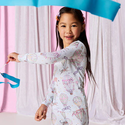 Young girl wearing a patterned onesie with colorful designs, holding blue ribbons against a white curtain background.