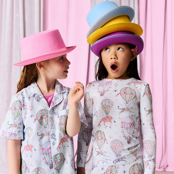 Two children wearing pajamas with a colorful hat stack on one of them against a pink curtain background.