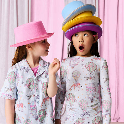 Two children wearing pajamas with a colorful hat stack on one of them against a pink curtain background.