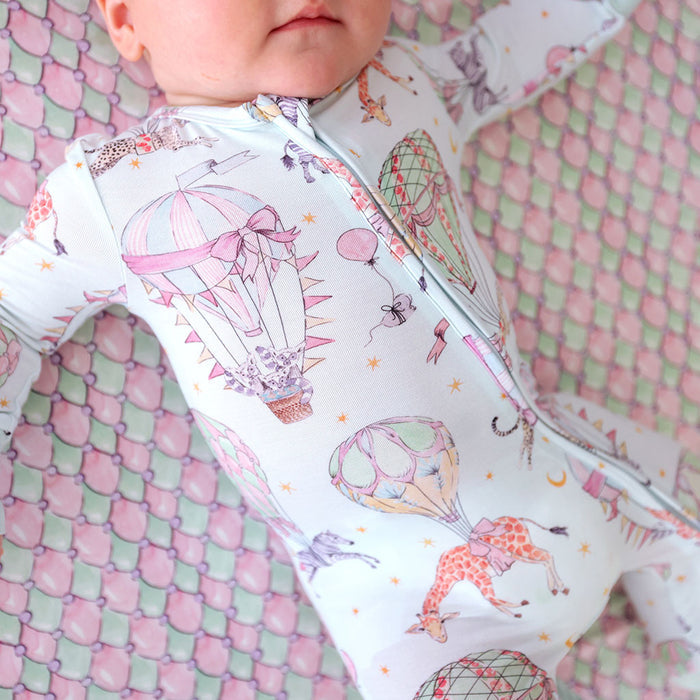 Baby wearing a colorful one piece with hot air balloons and giraffes on a pink and green checkered background.