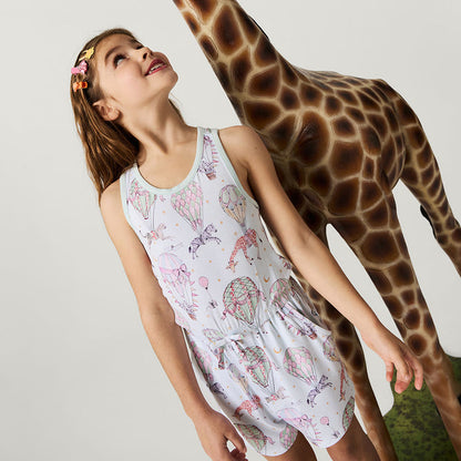 Young girl in a patterned romper standing next to a giraffe on a plain background