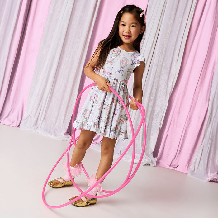 Young girl in a circus printed dress holding a pink hula hoop against a pink and gray curtain background