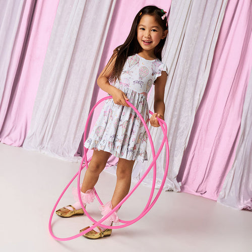 Young girl in a circus printed dress holding a pink hula hoop against a pink and gray curtain background