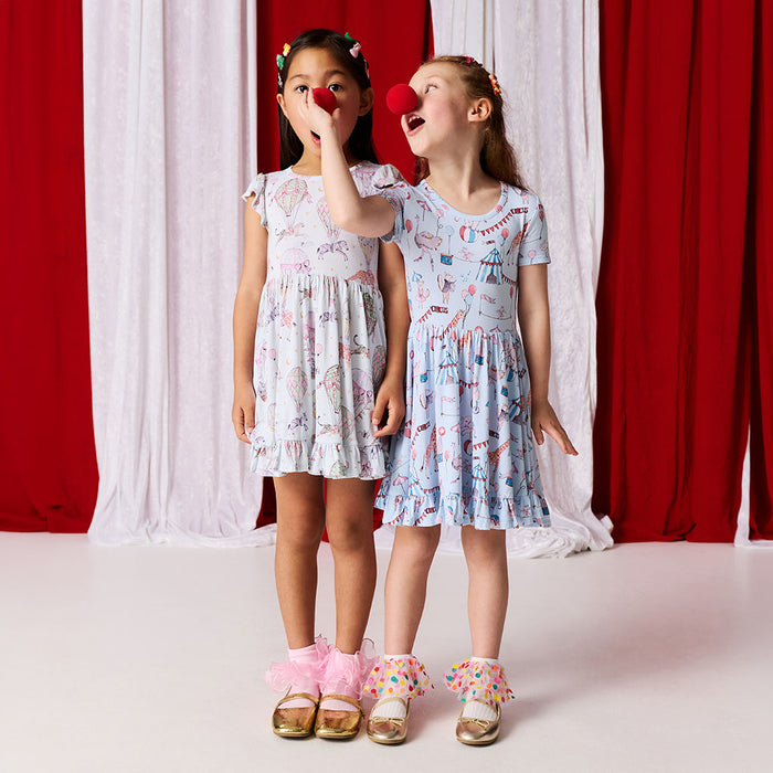 Two girls wearing matching circus printed dresses standing against a red and white curtain.