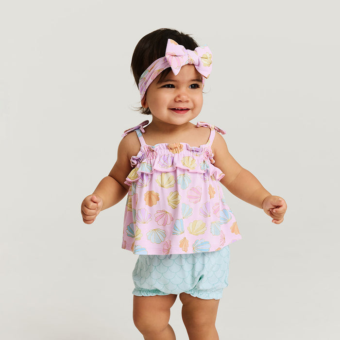 Baby wearing a colorful top and light blue shorts with a pink headband on a plain background
