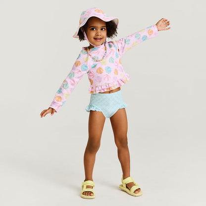 Child wearing a colorful shell printed rash guard, light blue shorts, and yellow sandals on a plain background