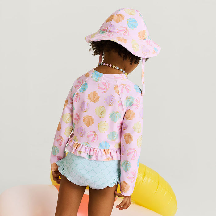 Child wearing a pink shell patterned sun hat and matching top with light blue shorts on a white background