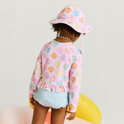 Child wearing a pink shell patterned sun hat and matching top with light blue shorts on a white background