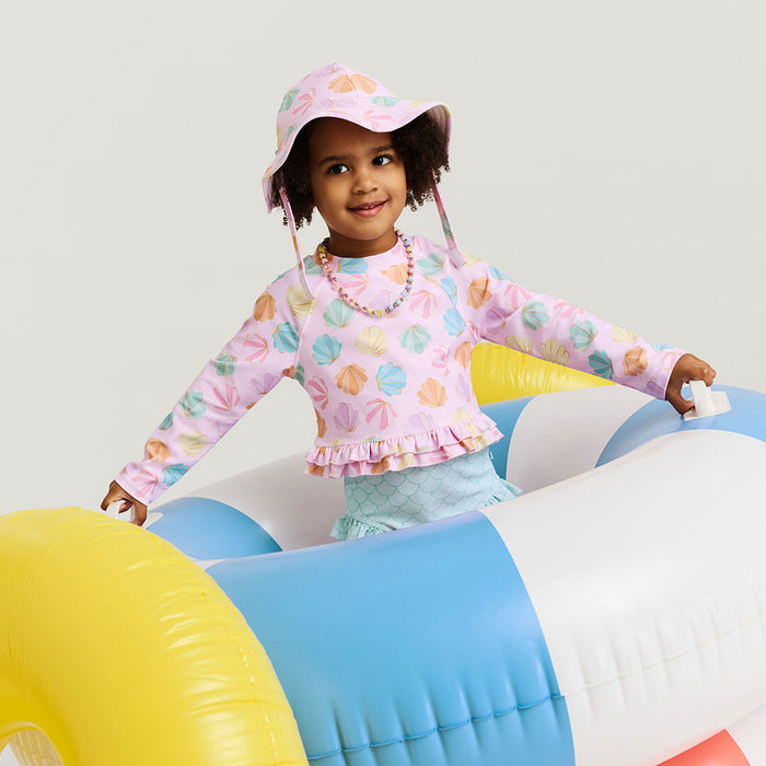 Child wearing a colorful shirt and hat, sitting on an inflatable toy with a plain background