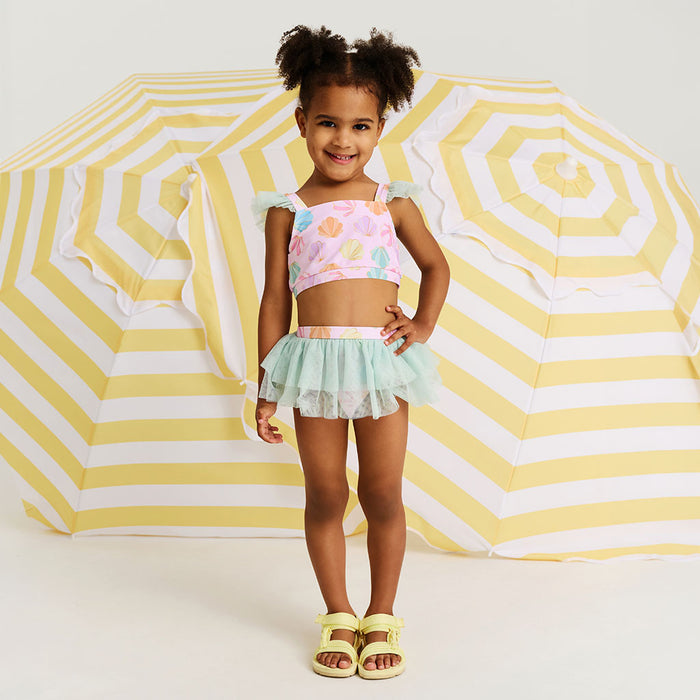 Young girl in a colorful two-piece swimsuit standing in front of a large yellow and white striped beach umbrella.