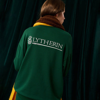 Person wearing a green sweater with 'SLYTHERIN' text and logo on the back against a dark curtain.
