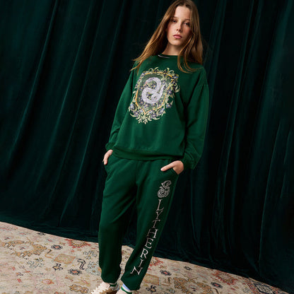 Person wearing a green jogger set with a Slytherin crest on a dark curtain background