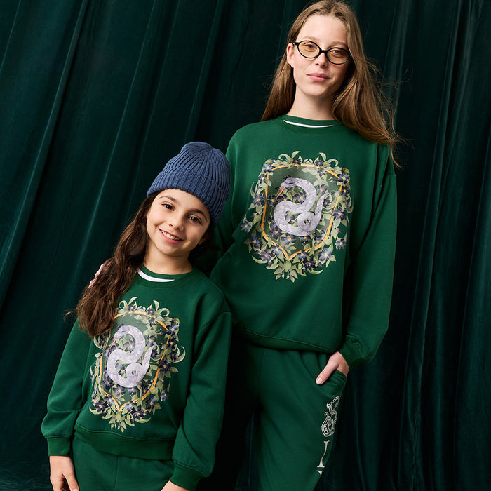 Two people wearing matching green sweatshirts with Slytherin crest against a dark green curtain.