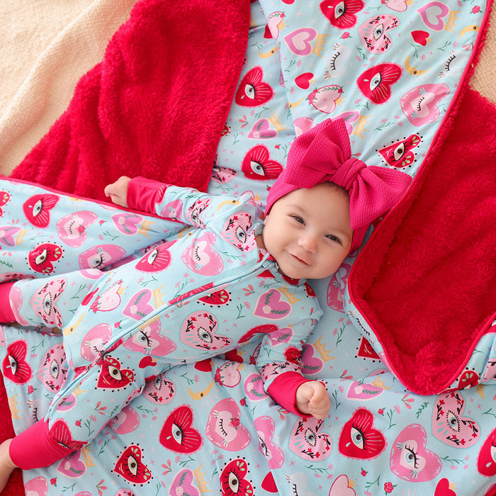 Baby in a blue heart printed one piece with a red bow headband on a matching blanket.